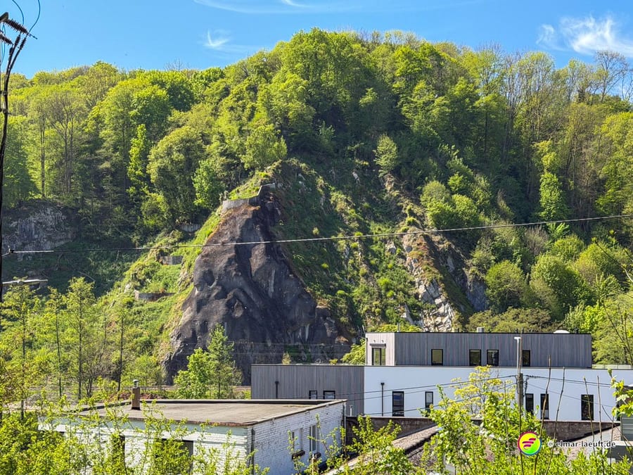 Bewaldeter Felsvorsprung über flachen Gewerbebauten im Tal der Ourthe.