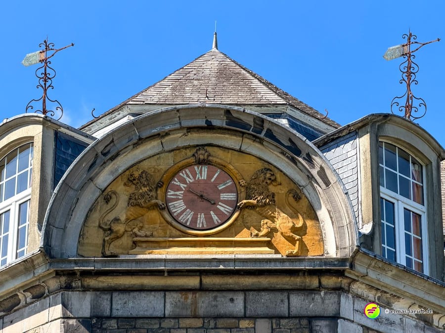 Giebel mit gemalter Uhr und goldenem Relief, links und rechts schmiedeeiserne Aufsätze.
