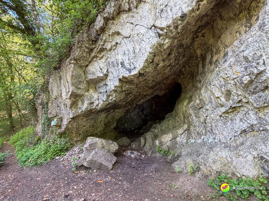 Höhleneingang in einer geschichteten Kalksteinwand direkt am Weg.