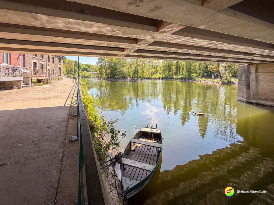 Unter einer Betonbrücke, am Ufer liegt ein hölzernes Boot im Wasser.