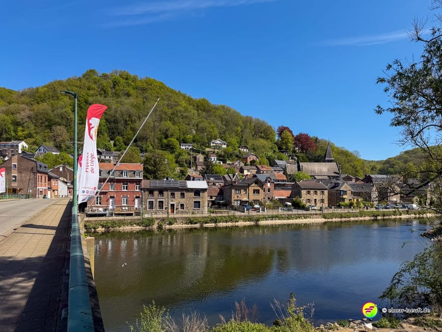 Von der Brücke auf den Fluss, am gegenüberliegenden Ufer Häuser und eine rote Fahne.