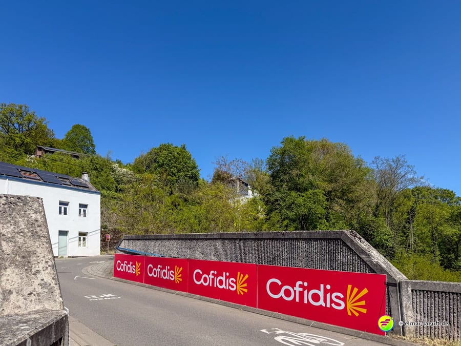Straße entlang einer langen Mauer mit knallrotem Cofidis-Schriftzug.