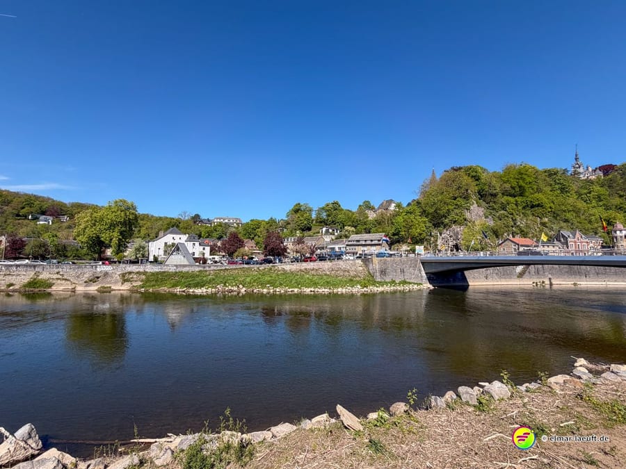 Kiesbank am Fluss, dahinter die Brücke, Häuser und ein Felsen.