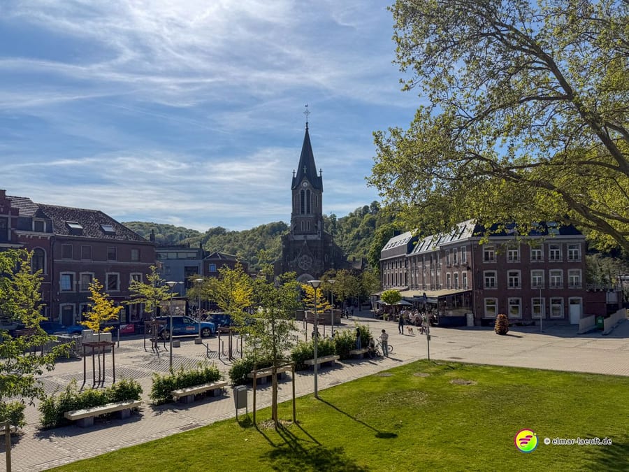 Platz im Ort mit Kirchturm, Bäumen und mehrstöckigen Häusern.