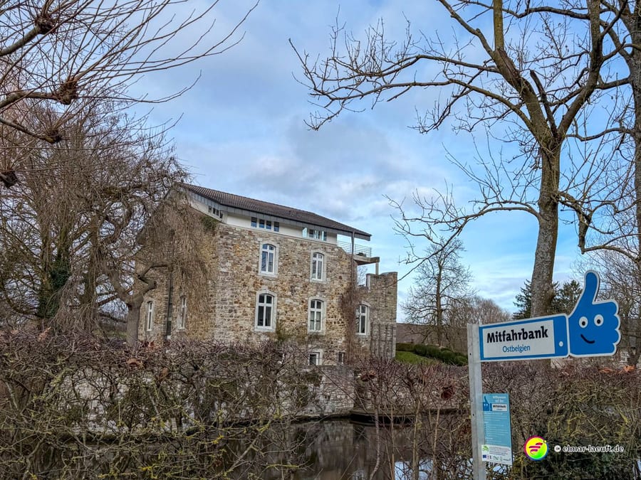 Altes Herrenhaus aus Naturstein mit Wassergraben und moderner Mitfahrbank. Das blaue Schild mit Daumen-Symbol zeigt 'Mitfahrbank' an – ein Ridesharing-Konzept für ländliche Gebiete.