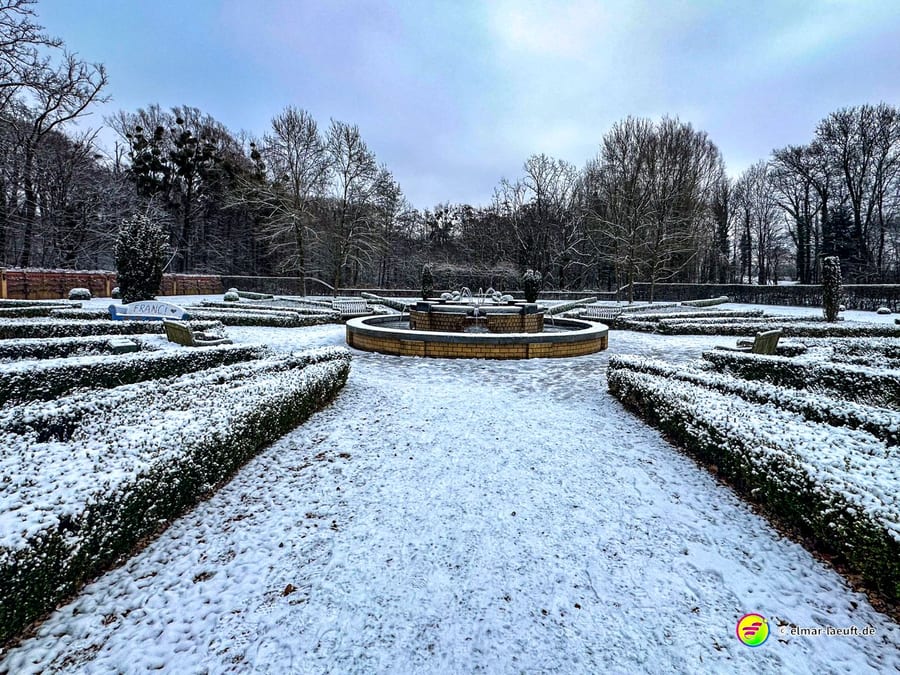 Laufen auf einem verschneiten, symmetrisch angelegten Gartenweg mit einem Brunnen in der Mitte und kahlen Bäumen im Hintergrund.