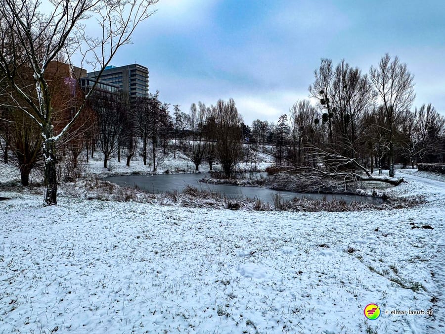 Laufen im winterlichen Heerlen entlang eines verschneiten Teichs mit kahlen Bäumen und Gebäuden im Hintergrund.