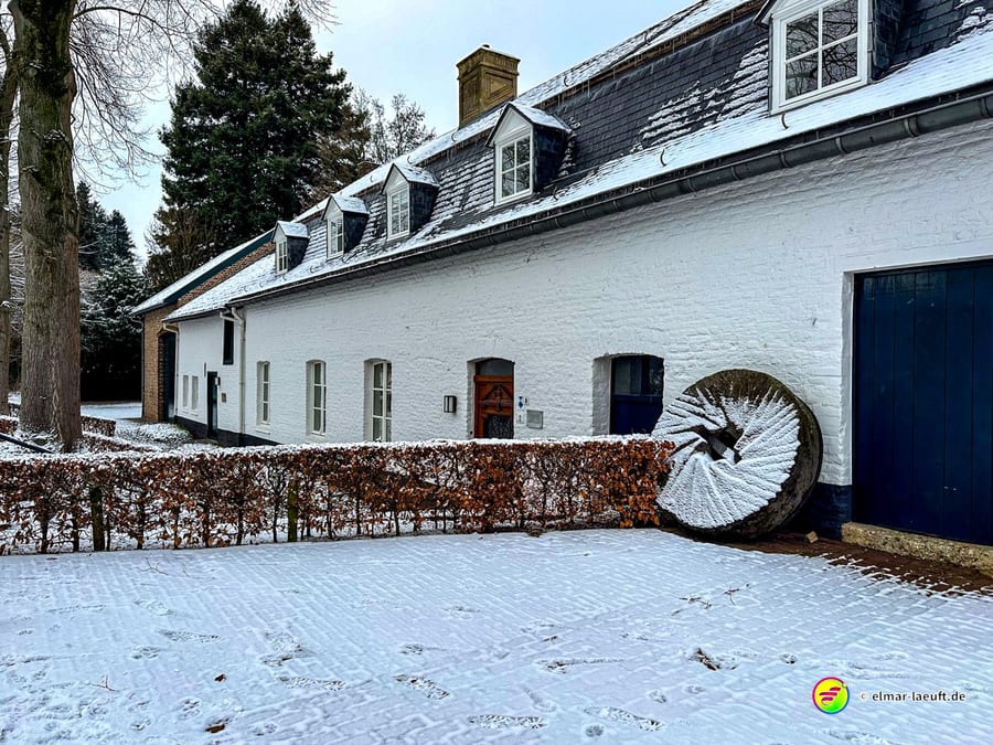 Laufen im verschneiten Heerlen vorbei an einem weißen Fachwerkhaus mit altem Mühlstein und kahlen Büschen.