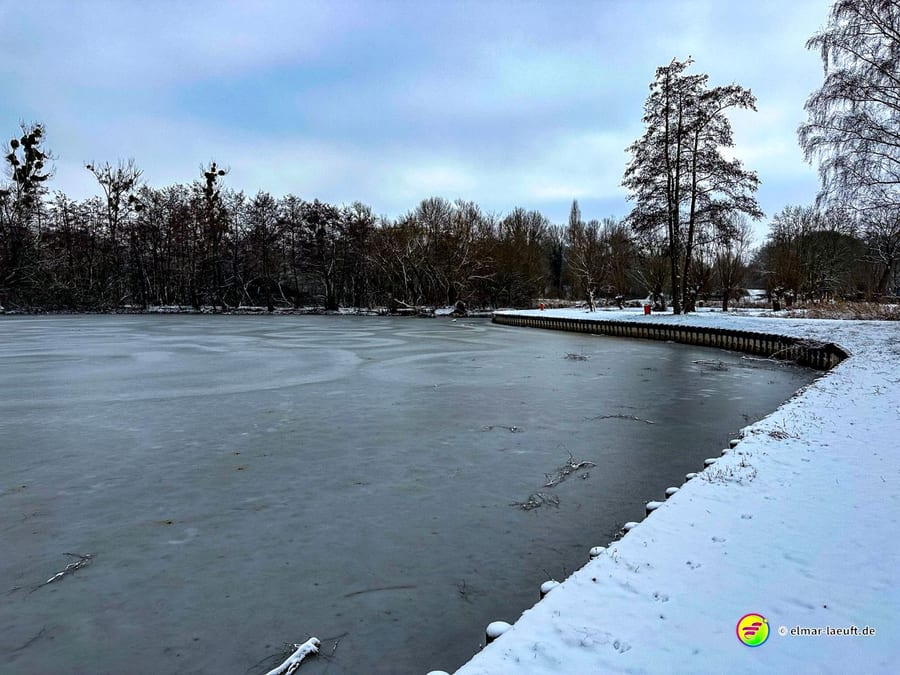 Laufen an einem zugefrorenen Seeufer im verschneiten Park von Heerlen mit kahlen Bäumen unter bewölktem Himmel.