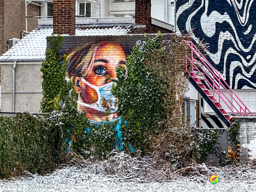 Laufen in Heerlen führt an einem mit Efeu bewachsenen Haus vorbei, dessen Wand ein großes Graffiti einer Frau mit Maske ziert, umgeben von leicht verschneiter urbaner Umgebung.