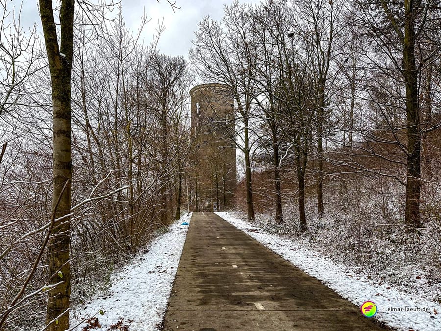Laufen auf einem schmalen, schneebedeckten Weg in einem winterlichen Wald mit einem alten, runden Turm im Hintergrund.