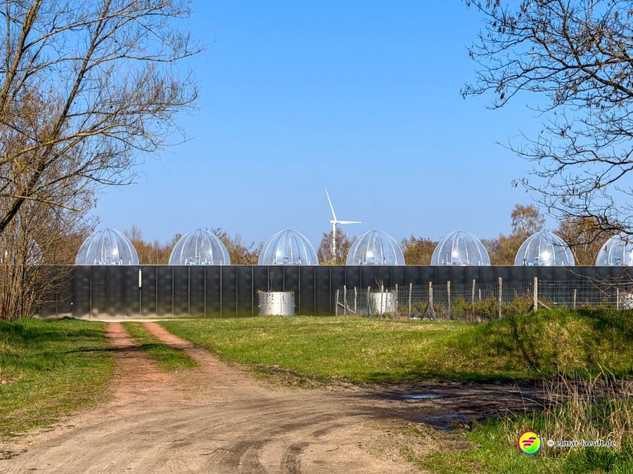 Laufen auf einem Feldweg in Maasmechelen mit Blick auf eine moderne Anlage mit gläsernen Kuppeln und einer Windkraftanlage im Hintergrund.