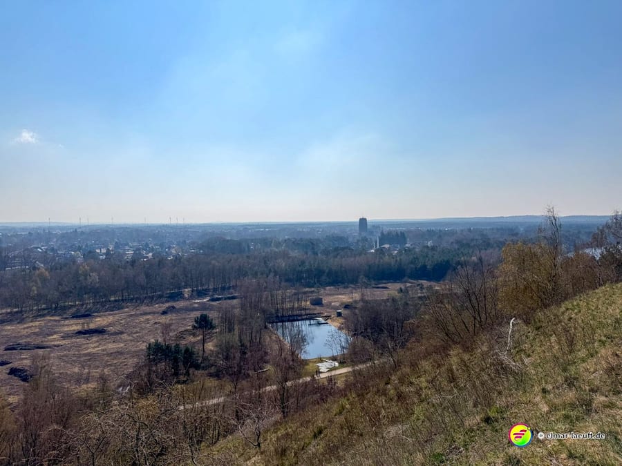 Blick beim Laufen in Maasmechelen von einem Hang auf eine offene Landschaft mit Bäumen, einem kleinen Teich und einer Stadt im Hintergrund.