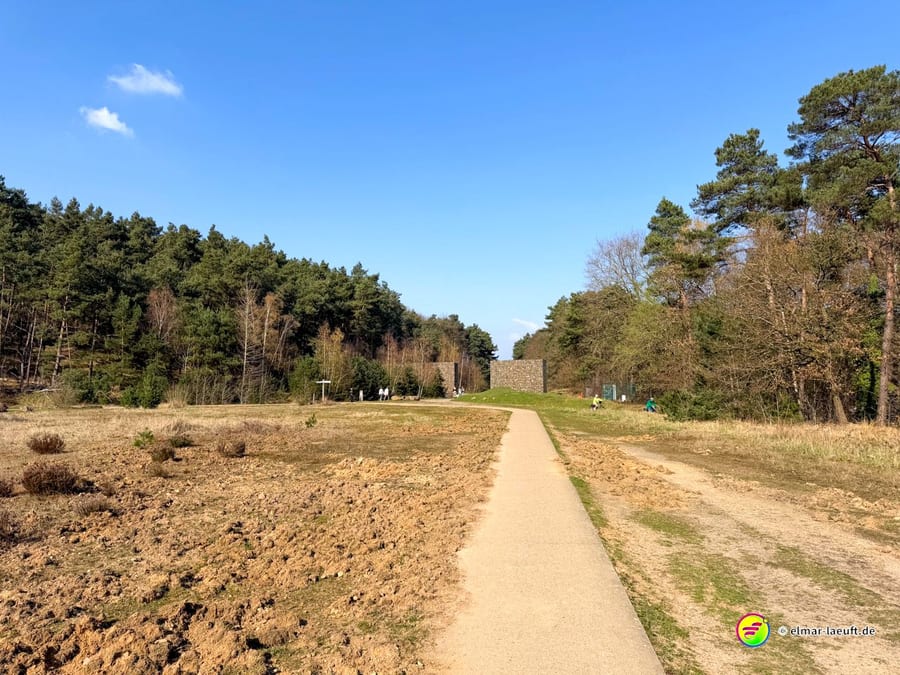 Laufen auf einem schmalen, befestigten Weg durch eine offene Landschaft mit Wald am Rand und einer markanten Steinmauer im Hintergrund bei klarem Himmel in Maasmechelen.