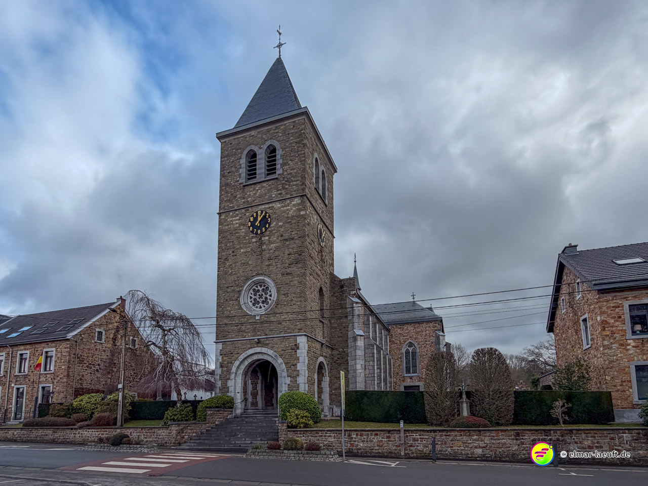 Steinkirche mit hohem Glockenturm und Uhr unter bewölktem Himmel in einem belgischen Dorf. Die Kirche zeigt romanische und neugotische Architekturelemente mit einem markanten quadratischen Turm und rundbogigem Haupteingang.