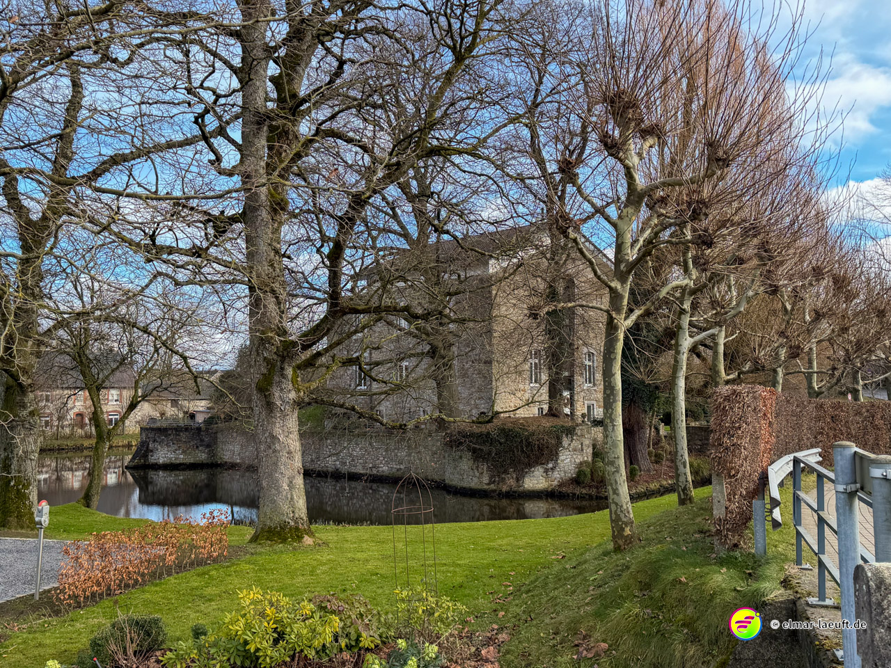 Historisches Wasserschloss mit Wassergraben umgeben von kahlen Bäumen im Vorfrühling. Die Burganlage aus Naturstein liegt malerisch hinter einer Steinbrücke in einer grünen Parklandschaft.
