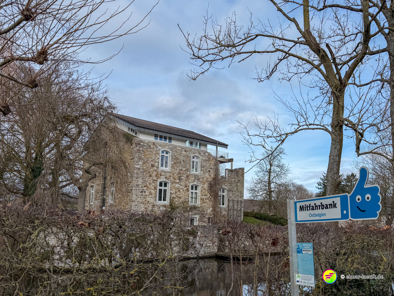 Altes Herrenhaus aus Naturstein mit Wassergraben und moderner Mitfahrbank. Das blaue Schild mit Daumen-Symbol zeigt 'Mitfahrbank' an – ein Ridesharing-Konzept für ländliche Gebiete.