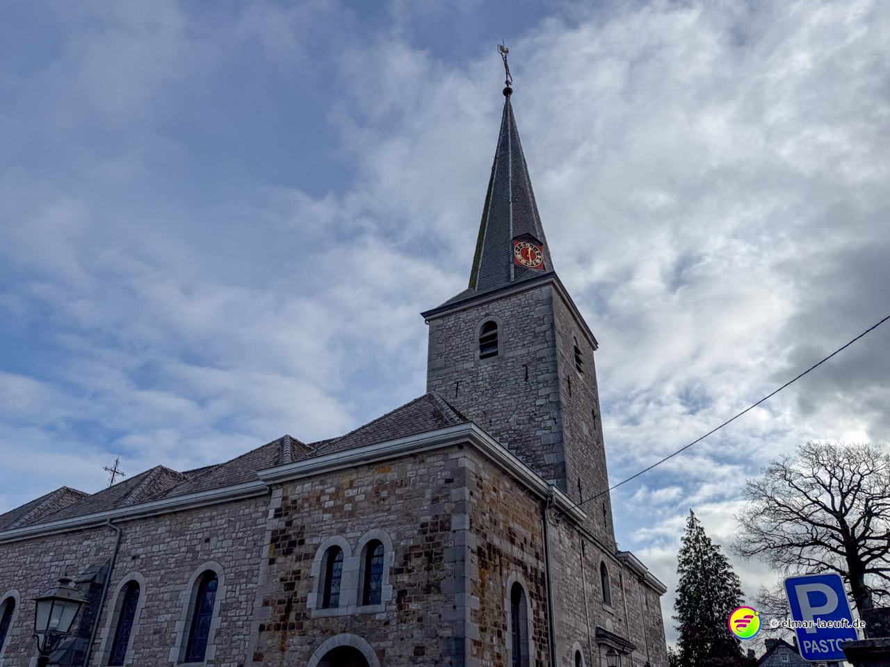 Romanische Dorfkirche mit grauem Steinturm und rotem Ziffernblatt unter dramatischem Wolkenhimmel. Der Kirchturm zeigt charakteristische Rundbogenfenster und einen spitzen Schieferhelm.