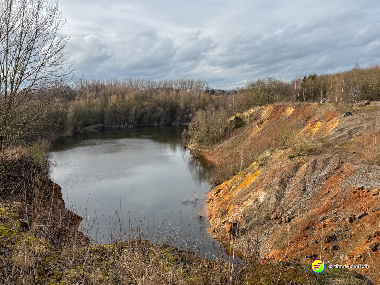 Aufgelassener Steinbruch mit See und charakteristischen orangefarbenen und roten Erdschichten. Die geologischen Formationen zeigen Zink- und Erzablagerungen der ehemaligen Bergbauregion.