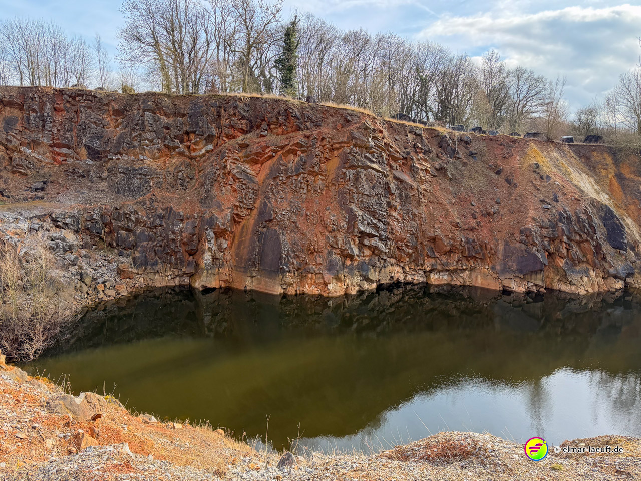 Dramatische mehrfarbige Gesteinsschichten am Rand eines Steinbruchsees mit orangefarbenen, roten und grauen Lagen. Die exponierten Erdschichten dokumentieren die mineralreiche Geologie der ehemaligen Zinkabbauregion.