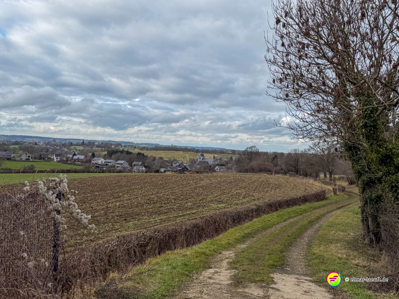 Unbefestigter Feldweg entlang einer Hecke mit Blick auf ein ländliches Dorf unter bewölktem Himmel. Der Wanderpfad führt durch die sanfte Hügellandschaft Ostbelgiens.