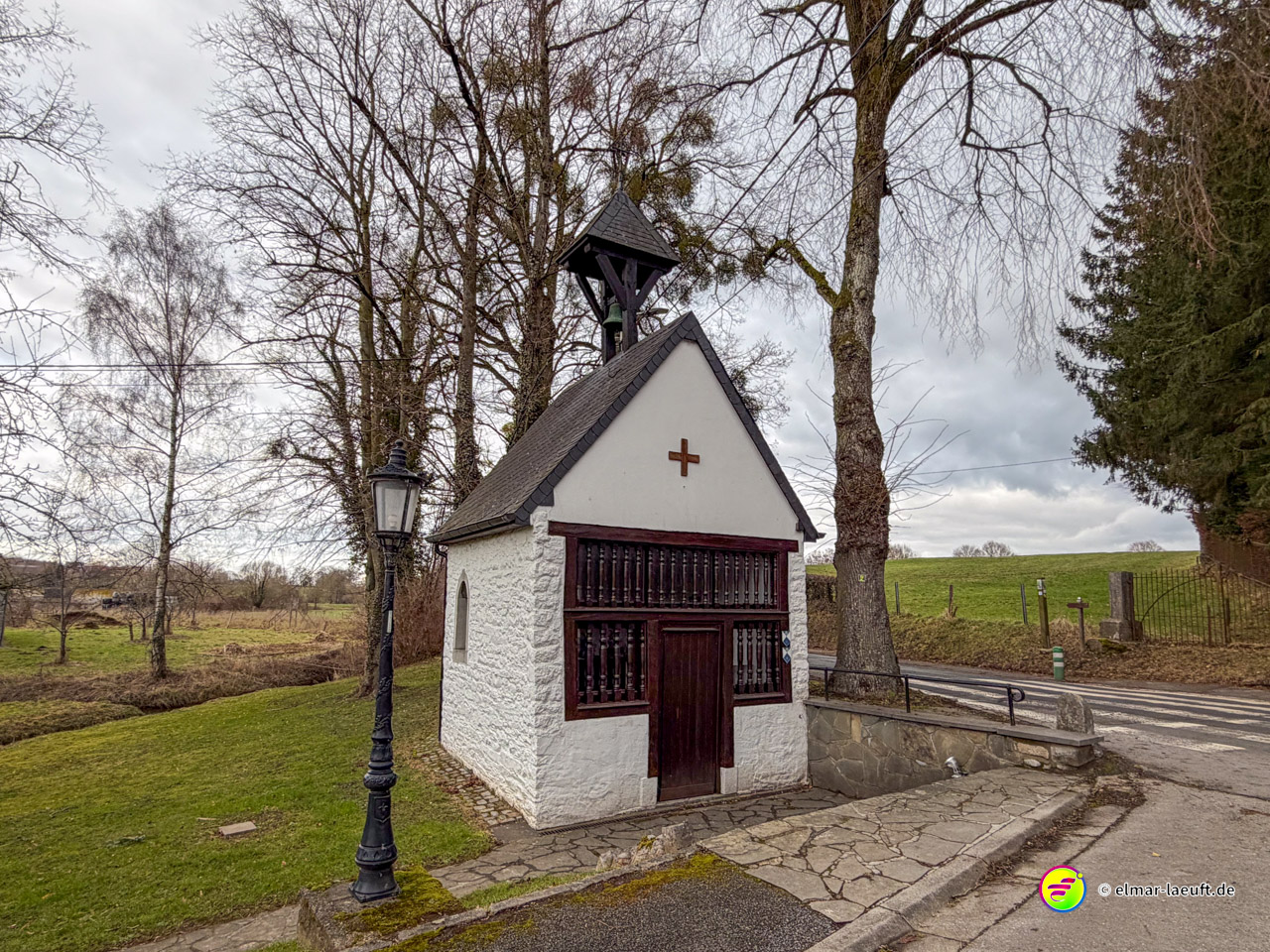 Kleine weiße Wegekapelle mit roter Holztür, Glockenturm und Kreuz. Die gepflegte Feldkapelle mit traditioneller Laterne dient als religiöser Rastplatz am Wanderweg.