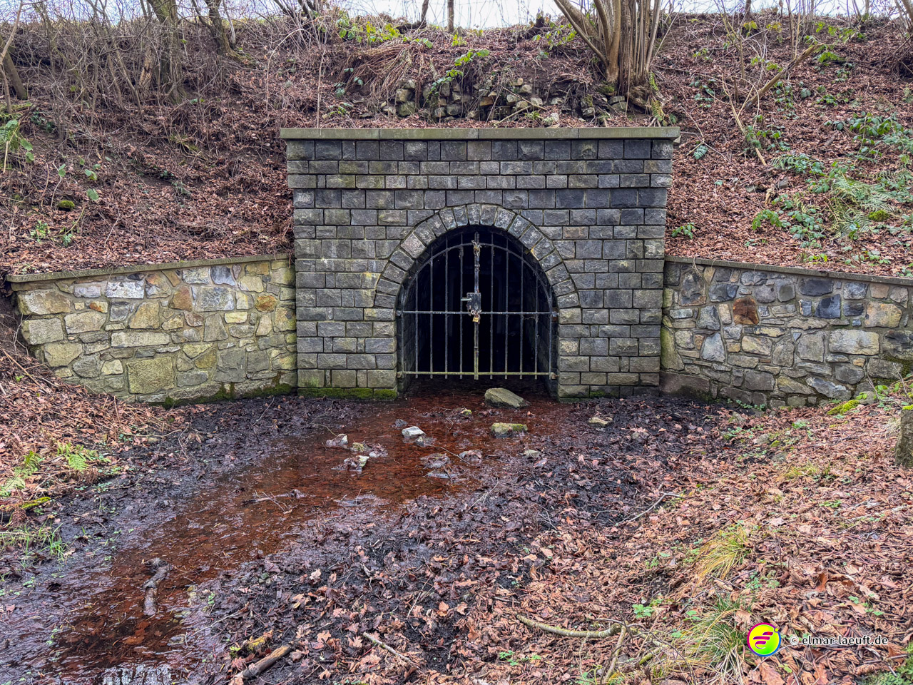 Verschlossener Stolleneingang in einen Hügel mit Eisengitter vor steinerner Portalfassade. Der alte Minenschacht zeigt rötliche Verfärbungen vom austretendem eisenhaltigem Wasser der Bergbauregion.