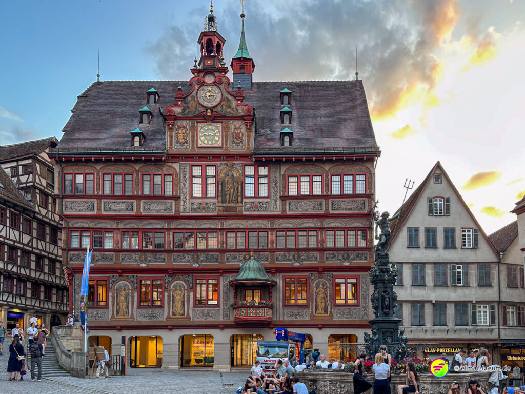 Laufen in Tübingen führt vorbei am historischen Rathaus mit kunstvoller Fassade und einem belebten Platz voller Menschen bei Abendlicht.