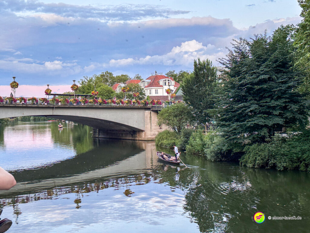 Laufen entlang eines ruhigen Flusses in Tübingen mit Blick auf eine blumengeschmückte Brücke und grüne Uferbereiche.