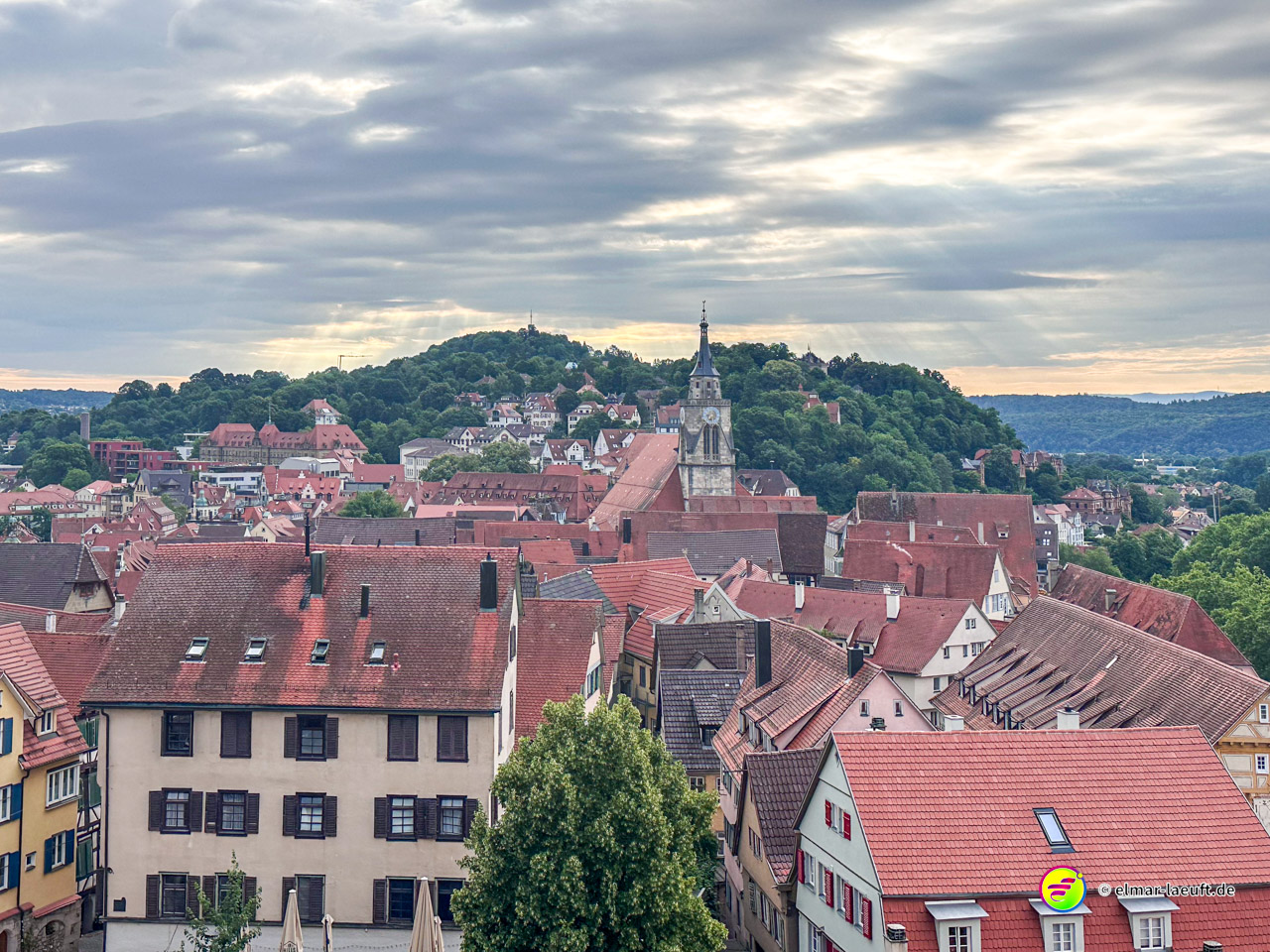 Blick beim Laufen über die Dächer von Tübingen mit der markanten Kirchturmspitze und bewaldeten Hügeln im Hintergrund unter einem bewölkten Himmel.