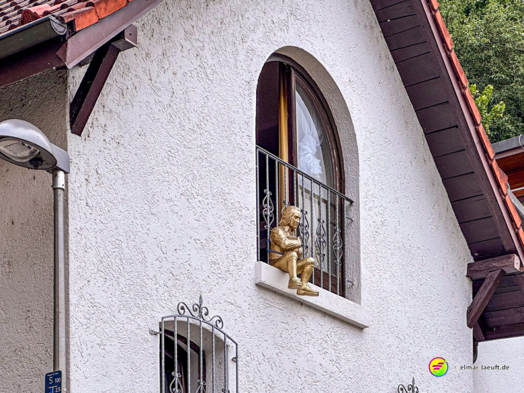 Beim Lauf durch Tübingen fällt eine goldene Statue auf, die auf einem Fensterbrett eines weißen Hauses mit rotem Dach sitzt.