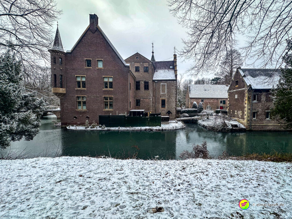 Laufen entlang eines verschneiten Ufers mit historischen Backsteinbauten und einem Wassergraben in einer ruhigen Winterlandschaft.