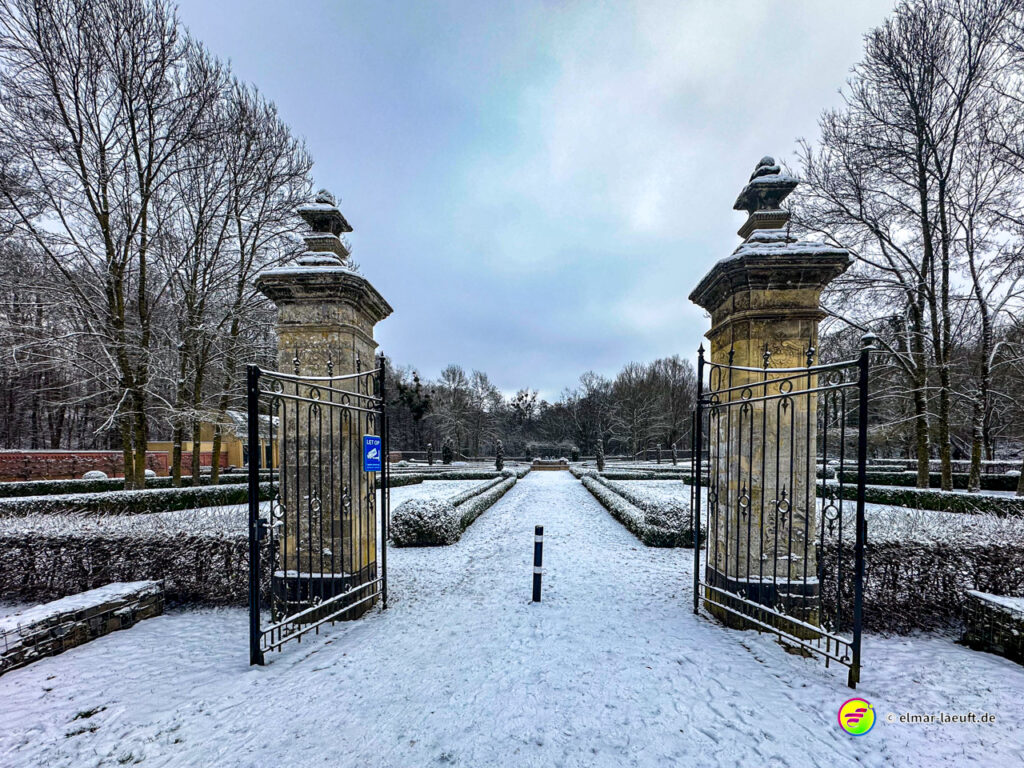 Laufen auf verschneitem Weg durch ein geöffnetes Tor mit steinernen Säulen in einem winterlichen Park mit kahlen Bäumen.