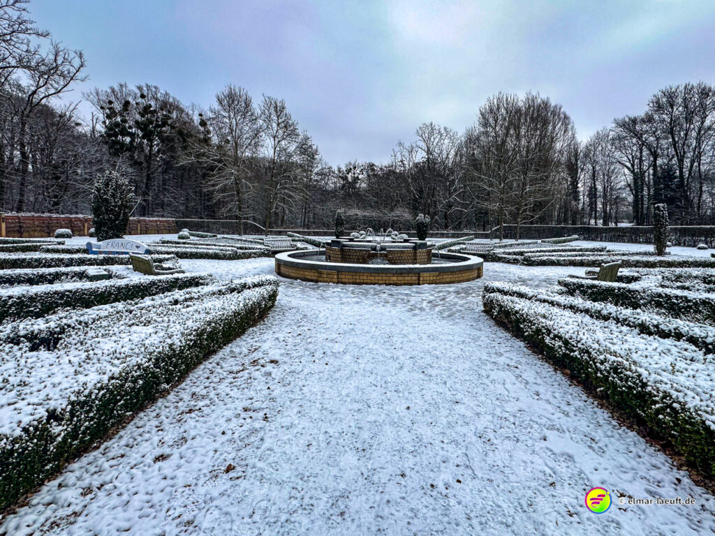 Laufen auf einem verschneiten, symmetrisch angelegten Gartenweg mit einem Brunnen in der Mitte und kahlen Bäumen im Hintergrund.