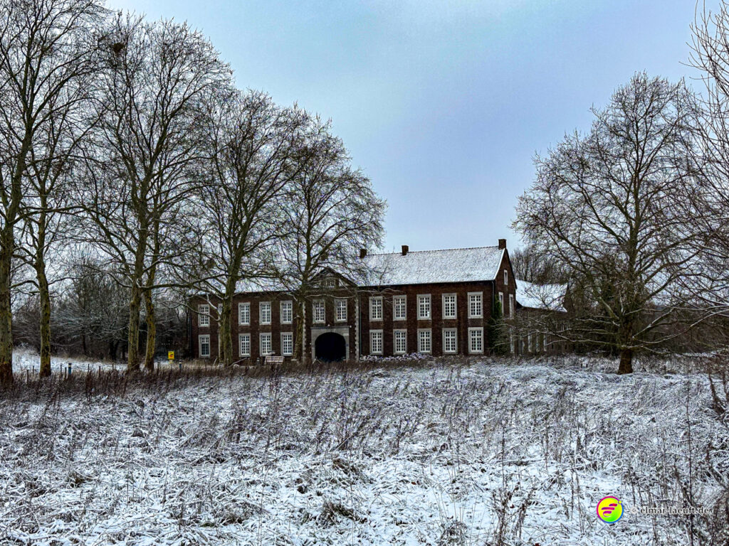 Laufen durch eine verschneite Wiesenlandschaft mit Blick auf ein altes, großes Backsteingebäude und kahle Bäume im Winter.