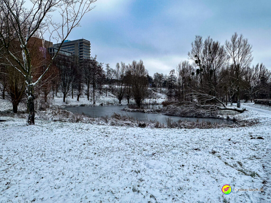 Laufen im winterlichen Heerlen entlang eines verschneiten Teichs mit kahlen Bäumen und Gebäuden im Hintergrund.