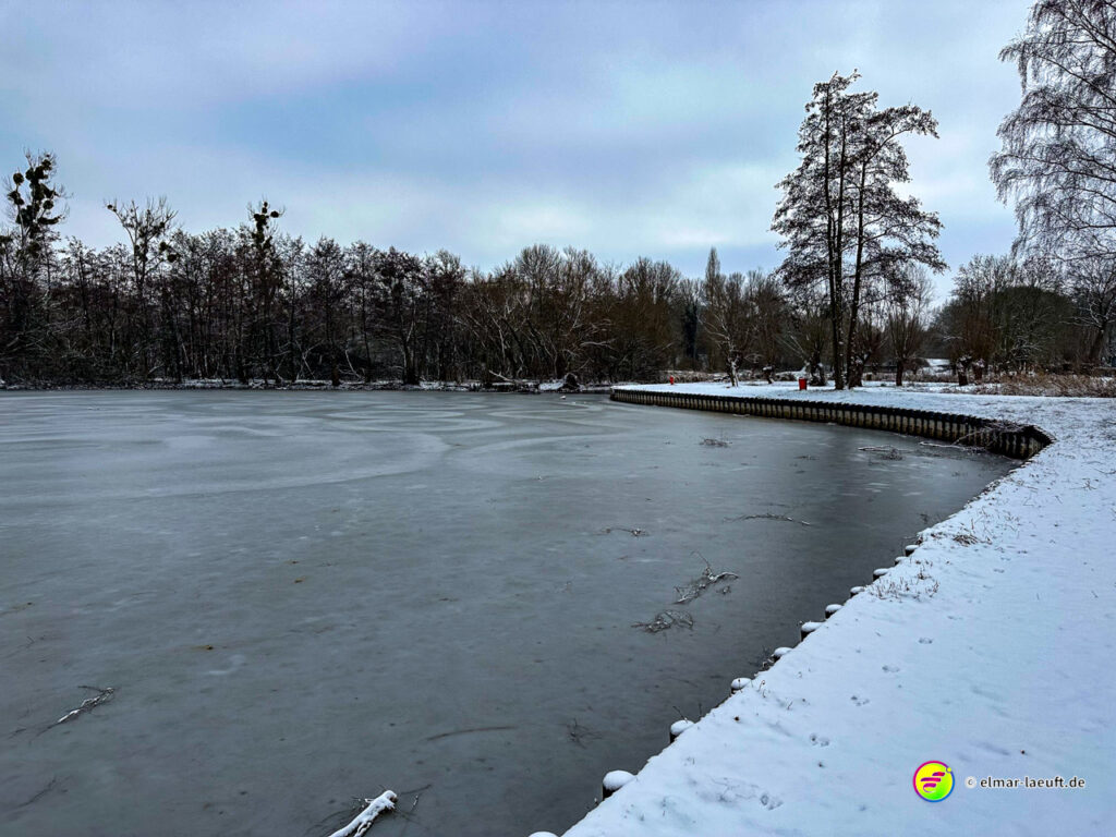 Laufen an einem zugefrorenen Seeufer im verschneiten Park von Heerlen mit kahlen Bäumen unter bewölktem Himmel.