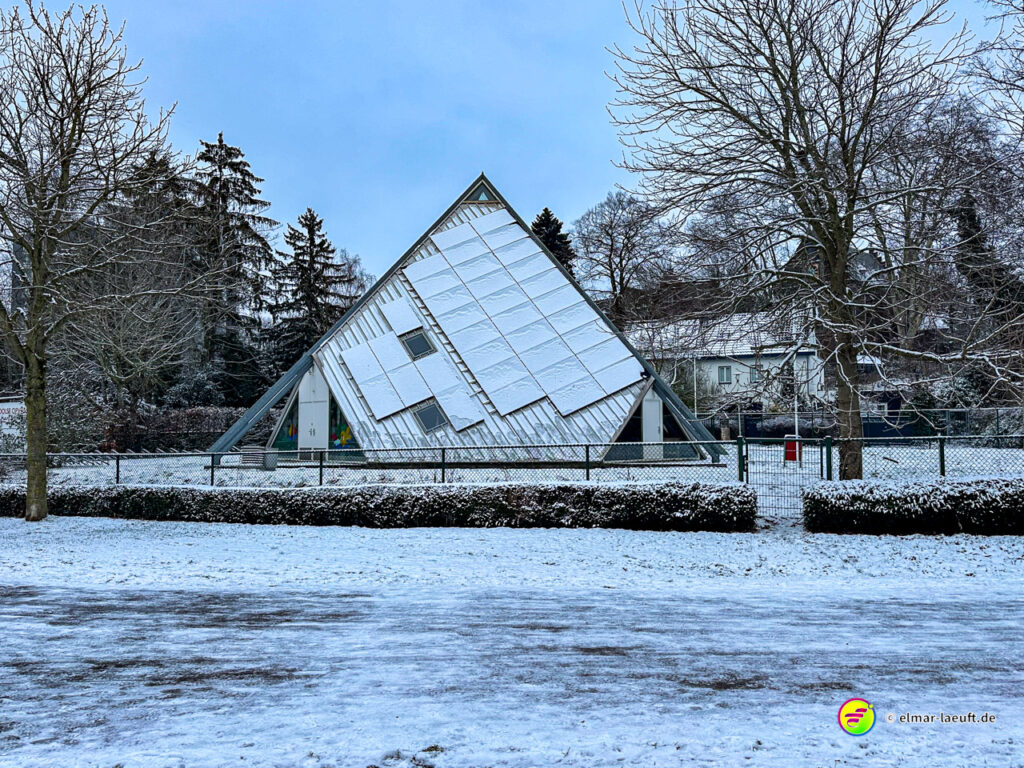 Laufen im winterlichen Heerlen vorbei an einem ungewöhnlich schräg stehenden, modernen Gebäude mit schneebedecktem Dach und kahlen Bäumen im Hintergrund.