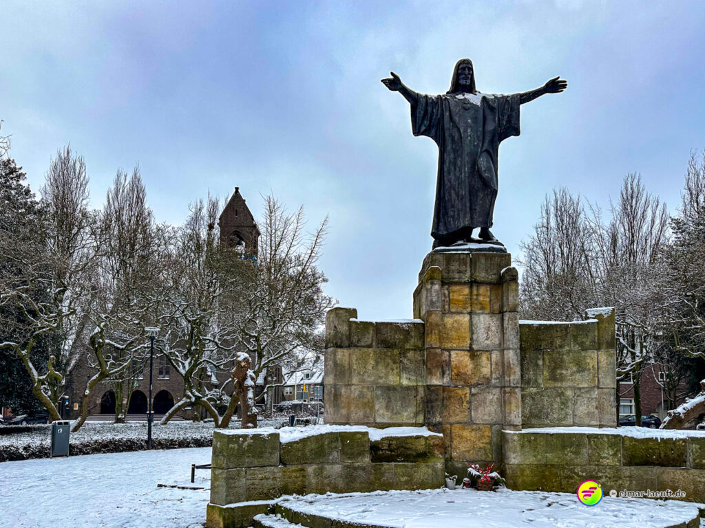 Laufen im winterlichen Heerlen vorbei an einer großen Christusstatue auf einem steinernen Sockel in einem verschneiten Park mit kahlen Bäumen und einer Kirche im Hintergrund.