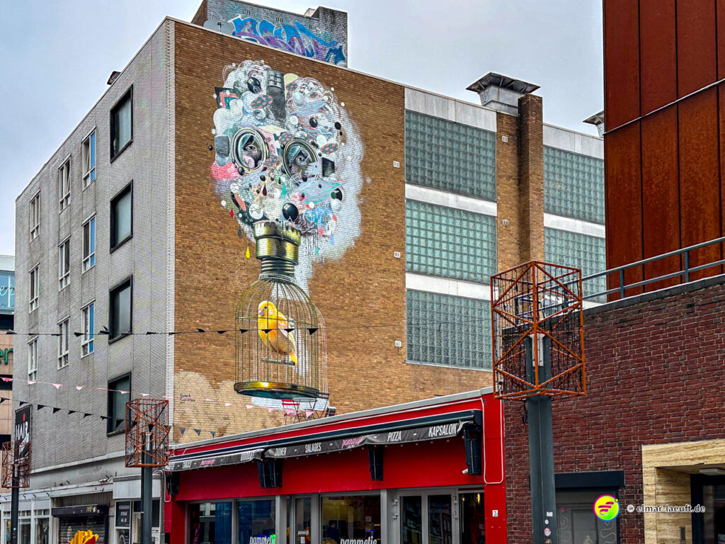 Laufen durch die Stadt Heerlen mit Blick auf ein großes Wandbild eines Käfigs mit einem gelben Vogel an einer Hauswand.