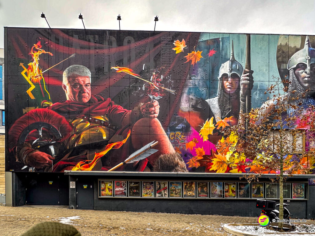 Laufen in Heerlen vorbei an einer großen, farbenfrohen Wandmalerei mit römischen Soldaten und herabfallenden Herbstblättern in einer urbanen Umgebung.