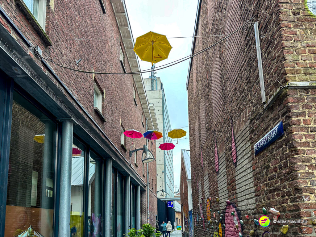Laufen durch eine enge Gasse in Heerlen mit bunten Regenschirmen, die über dem Kopf hängen, und Backsteinmauern auf beiden Seiten.