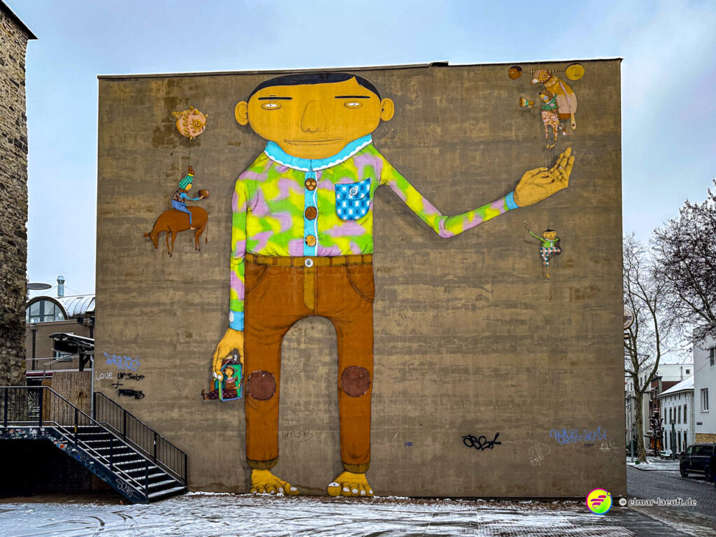 Laufen in Heerlen vorbei an einer großen bunten Wandmalerei eines stilisierten Menschen auf einer Hauswand in einer urbanen Umgebung mit leicht verschneitem Boden.