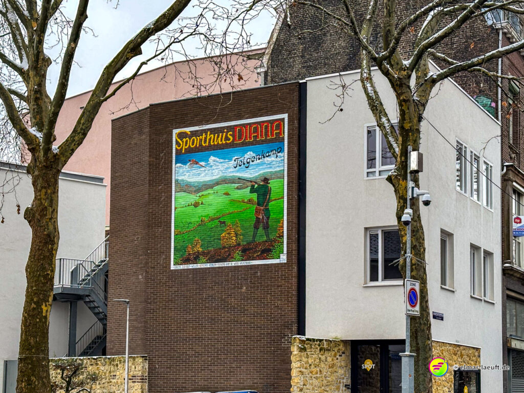 Laufen in Heerlen an einem urbanen Abschnitt mit einem Wandbild eines Jägers auf einem Gebäude zwischen kahlen Bäumen.