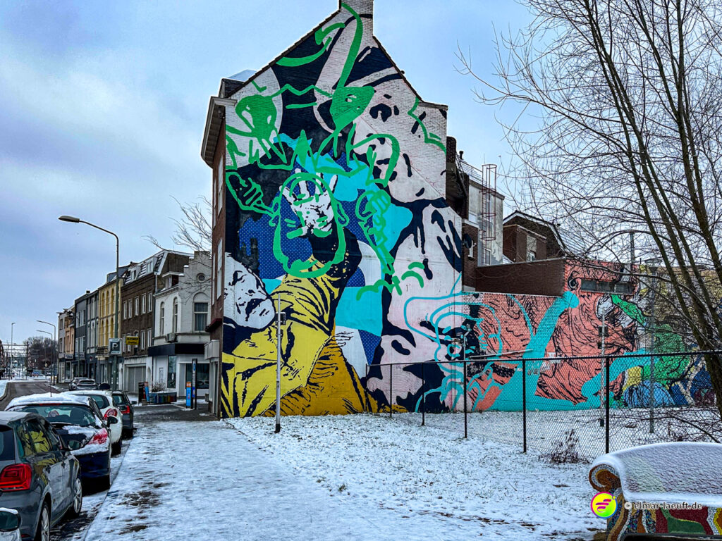 Laufen auf einem schneebedeckten Bürgersteig in Heerlen mit buntem Wandgemälde an einem Haus und Reihenhäusern entlang der Straße.