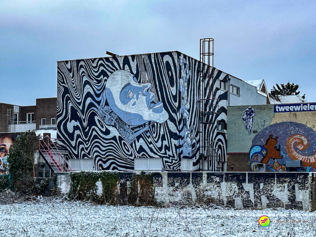 Laufen in Heerlen vorbei an einem Gebäude mit auffälligem schwarz-weißem Wandgemälde und leicht verschneitem Boden.