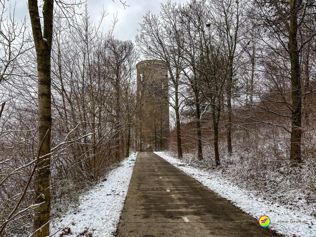 Laufen auf einem schmalen, schneebedeckten Weg in einem winterlichen Wald mit einem alten, runden Turm im Hintergrund.