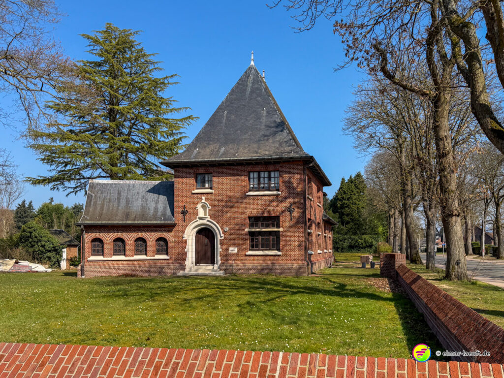 Laufen in Maasmechelen vorbei an einem historischen Backsteinhaus mit spitzem Dach und umgeben von Bäumen bei klarem Himmel.