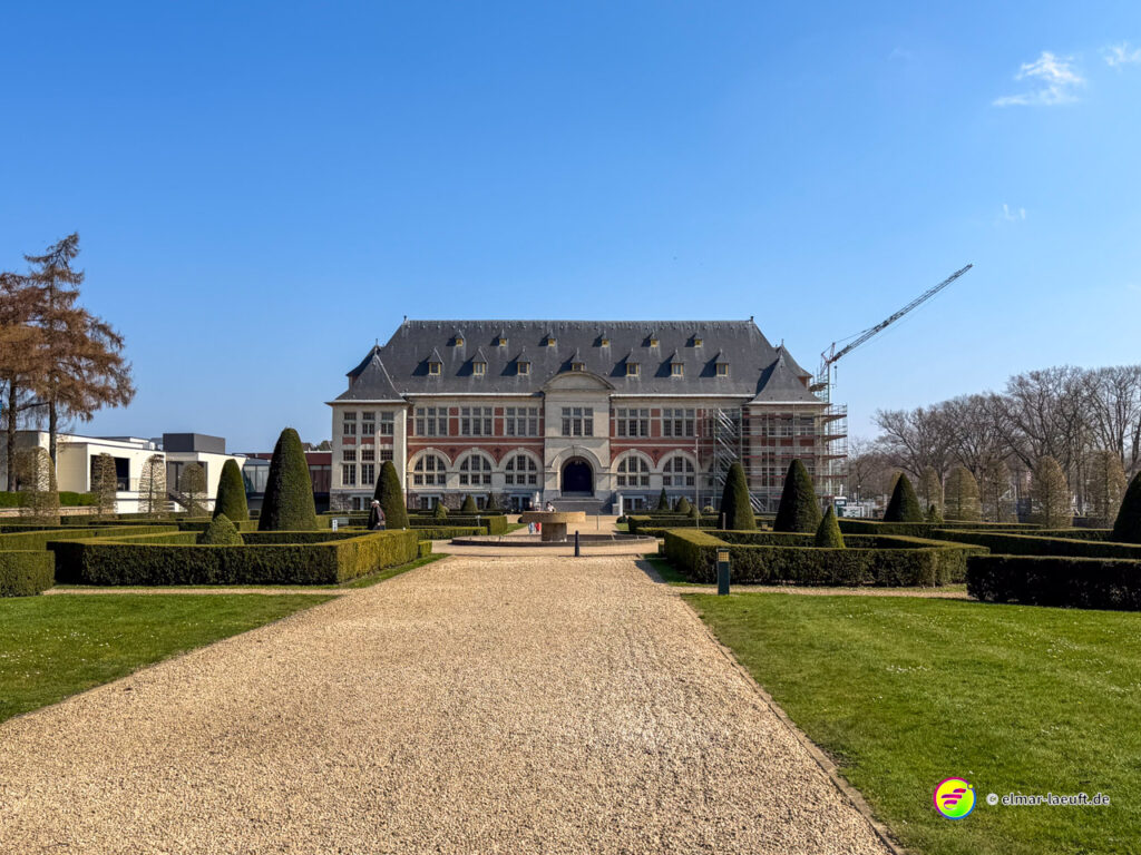 Laufen auf einem breiten Kiesweg durch einen gepflegten Garten mit symmetrisch geschnittenen Hecken und Blick auf ein großes historisches Gebäude in Maasmechelen.