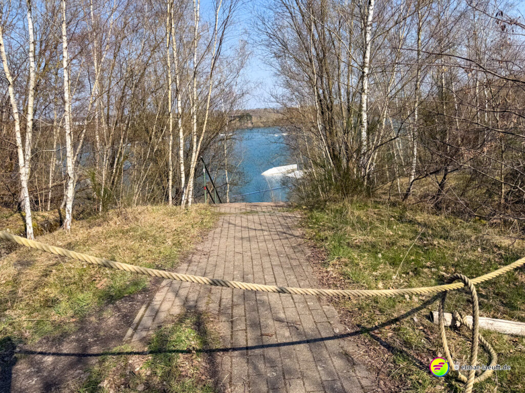Lauf auf einem gepflasterten Weg in Maasmechelen, der durch kahle Bäume zu einem See führt, mit einem dicken Seil als Sperre quer über dem Pfad.